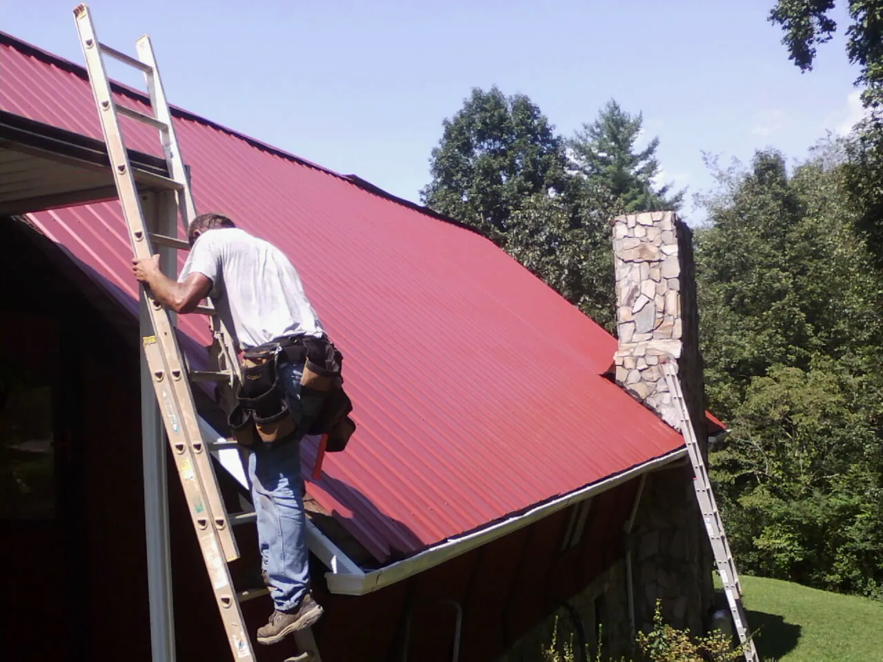 Roofer on a ladder beside a red metal roof and stone chimney — Soffit & Fascia Repair by Roofing Brothers in Lake Villa
